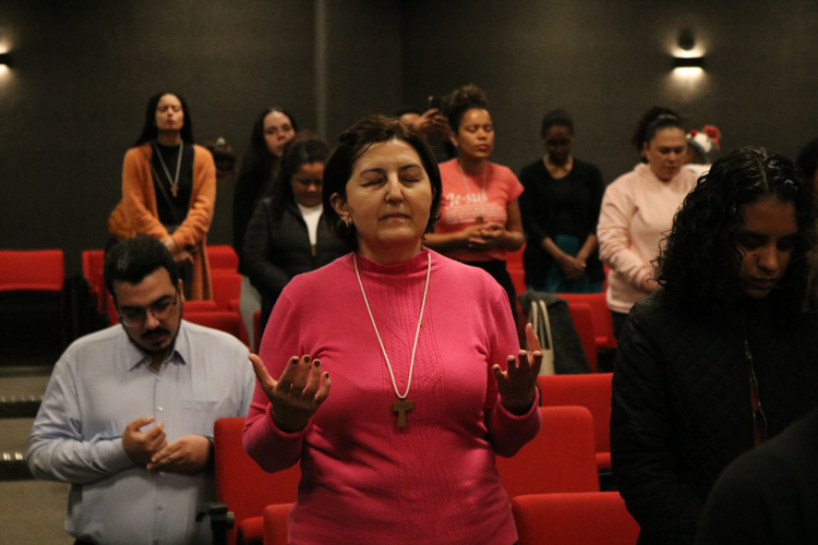 Mulher de blusa rosa e terço no pescoço ora com olhos fechados e mãos erguidas, entre outros fiéis concentrados em oração.