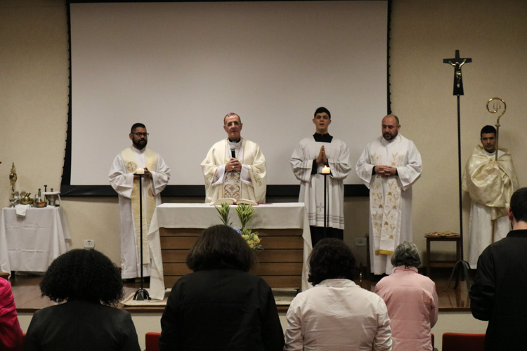 Vista ampla do altar com Dom Carlos Lema e outros sacerdotes celebrando a missa, enquanto fiéis rezam nas primeiras fileiras do auditório.