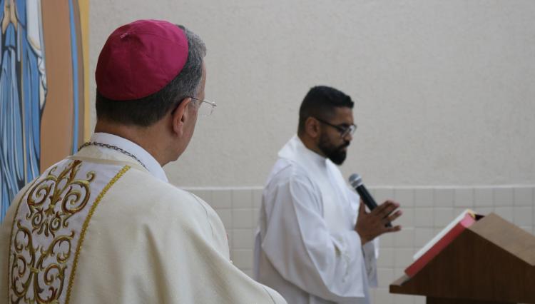 Dom Carlos Lema, visto de costas, observa enquanto o Padre Rodrigo Felipe, segurando um microfone, conduz uma leitura durante a Missa de Abertura do Ano Letivo na capela do Centro Universitário Assunção