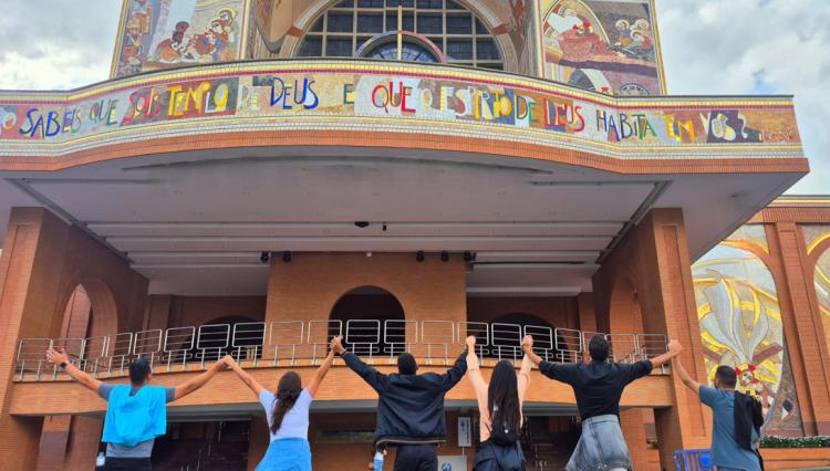 Grupo de sete pessoas de mãos dadas, de costas para a câmera, em frente à Basílica de Nossa Senhora Aparecida, em Aparecida (SP).