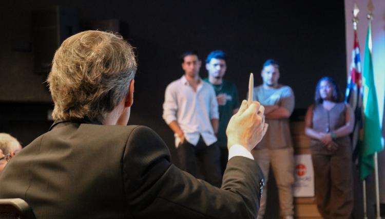 Integrante da banca avaliadora, de costas, segura uma caneta enquanto um grupo de quatro estudantes apresenta um projeto no palco durante o evento Tubarões da Administração 2025, no auditório do Centro Universitário Assunção. Ao fundo, bandeiras do Brasil e do estado de São Paulo